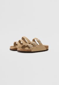 Beige suede sandals with three adjustable straps and silver buckles, set on a cork footbed and textured brown rubber outsole.