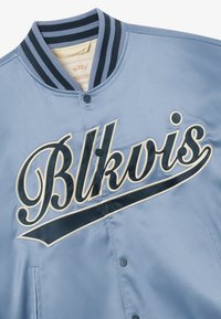 Veste bomber en satin bleu clair avec col rayé noir et blanc, boutons-pression, et le mot « Blkvis » brodé en gros caractères sur la poitrine.
