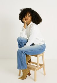 Camisa blanca de botones con detalles de encaje, jeans acampanados azules y botines de tacón color beige, sentada en un taburete de madera contra un fondo blanco.