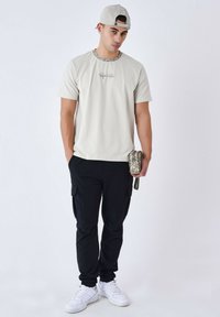 Camiseta de manga corta color gris claro con cuello estampado, combinada con pantalones cargo negros y zapatillas blancas; sosteniendo un pouch con estampado de camuflaje.