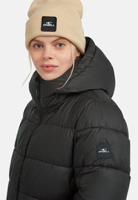 Zwarte pufferjas met gewatteerd ontwerp en hoge kraag. Gecombineerd met een beige gebreide beanie met een zwart logopatch.
