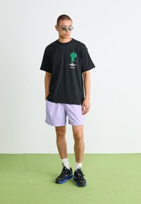 Mann iført svart t-skjorte med grønn kaktusprint, lys lilla shorts, hvite sokker, svarte og blå joggesko, og små solbriller som står på grønt gulv.