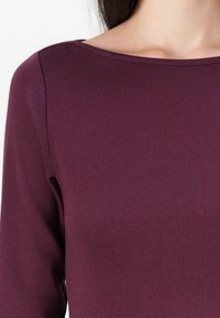 Nærbilde av en person som har på seg en langermet, crew neck, mørk lilla skjorte, som viser nakken og øvre del av torsoen mot en ensfarget bakgrunn.