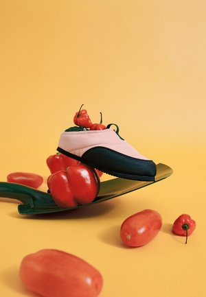 Pink og sort sneakers med chilipebre indeni, balancerende på en grøn skovl med røde peber spredt på en gul baggrund.