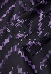 Tessuto nero con un motivo geometrico di linee e blocchi viola. Il design presenta larghezze e angoli variabili, creando un effetto testa.