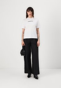 White short-sleeve T-shirt with black text, paired with high-waisted black wide-leg trousers and black heeled ankle boots. Holding a black bag.