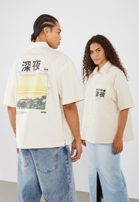 Cămăși bej cu mânecă scurtă, având un grafic pe spate care ilustrează o scenă de apus, cu text negru și accente. Modelele poartă pantaloni din denim casual.