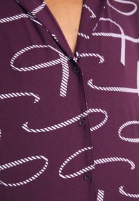 Camisa de color púrpura oscuro con un patrón de cuerda blanca, abotonada con dos botones negros en el cuello, hecha de una tela suave.