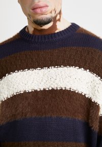 Brun, marinblå och krämfärgad randig tröja med ett texturerat stickmönster och ett brett, fluffigt krämfärgat band över bröstet. Rund halsringning.