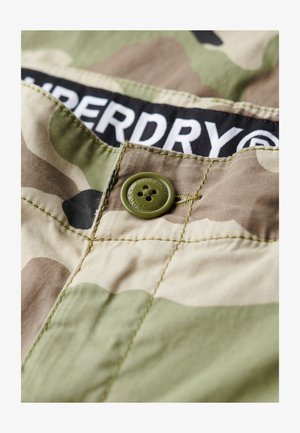 Tkanina w kamuflażowym wzorze w odcieniach zieleni i brązu, z zielonym guzikiem i czarną metką z białym napisem "SUPERDRY."