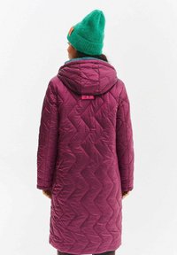 Fialový prošívaný dlouhý kabát s kapucí, zdobený cikcak vzorem. Na hlavě je zelená pletená čepice, která přidává kontrastní barvu.