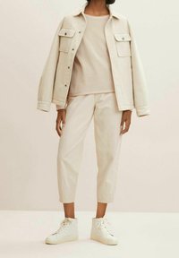 Persoon met crèmekleurige cropped broek, mesh top, oversized jas en witte hoge sneakers tegen een neutrale achtergrond.