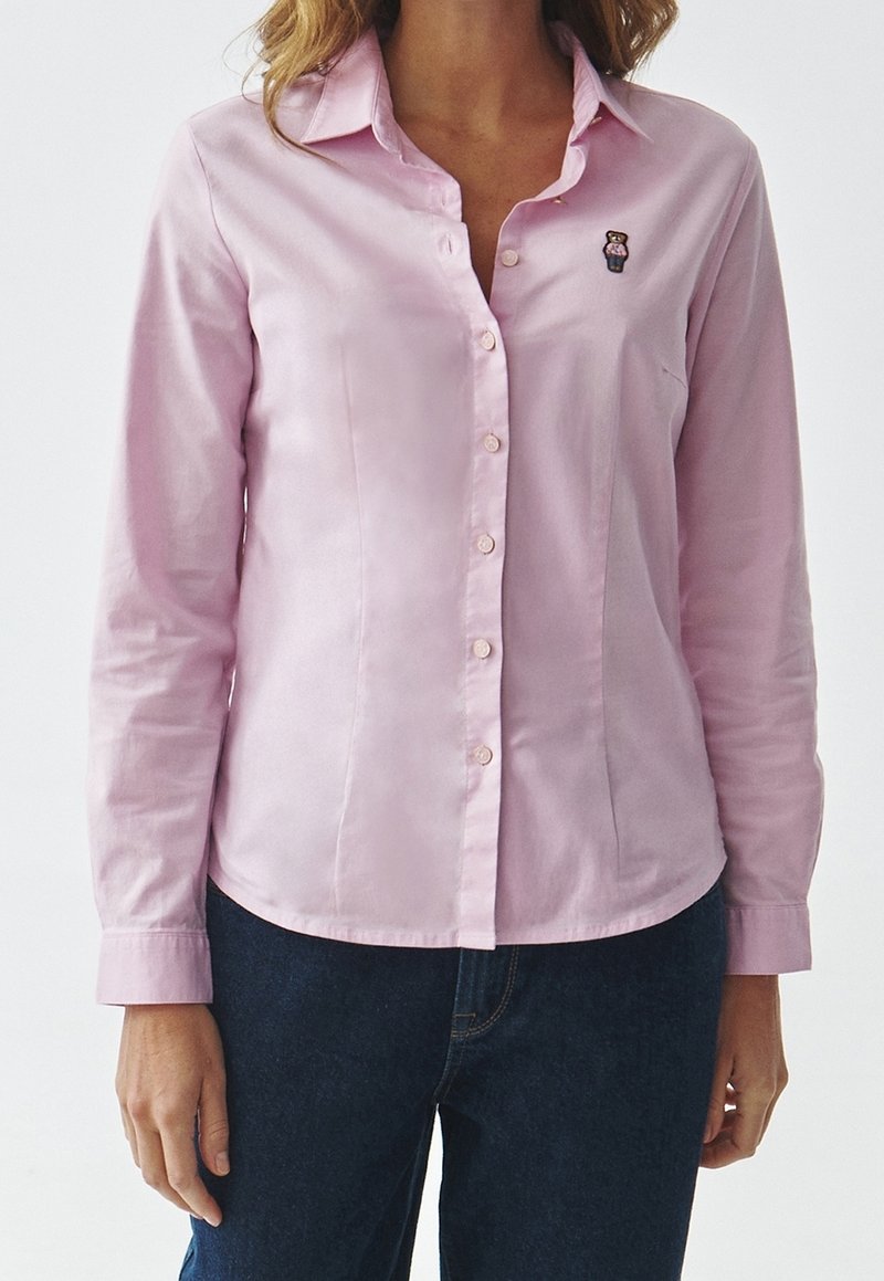 Femme portant une chemise boutonnée rose clair avec un petit ours brodé sur le côté gauche de la poitrine, assortie à un jean bleu foncé.