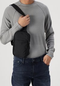 Svart axelväska i slitstarkt tyg, med en dragkedjeförsedd framficka, rektangulär form och justerbar rem, bärs över en grå tröja.