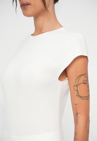 T-shirt blanc ajusté avec des manches courtes en forme de cap, fabriqué dans un tissu lisse. Présente un col rond et un tatouage sur le bras supérieur.