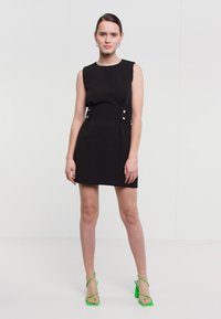 Γυναίκα σε μαύρο μίνι φόρεμα χωρίς μανίκια και φωτεινές πράσινες σαμπό με λουριά, που στέκεται σε απλό λευκό φόντο.