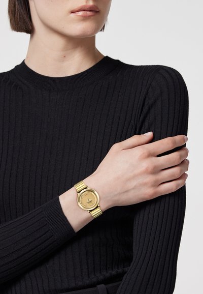 Femme portant un haut noir côtelé à manches longues, montrant une montre-bracelet en or avec un bracelet tissé au poignet gauche, la main reposant sur le bras droit.
