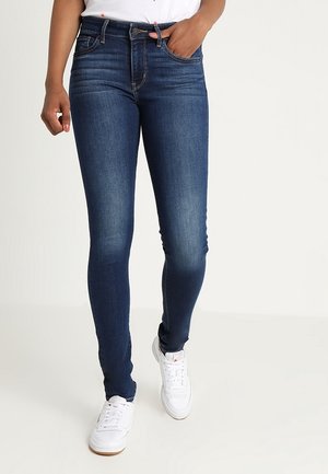 Kvinde iført mørkeblå skinny jeans og hvide sneakers, stående med den ene hånd i forlommerne mod en ensfarvet baggrund.