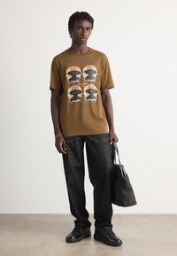 Junger Mann in einem braunen Grafik-T-Shirt, schwarzen Jeans und schwarzen Schuhen, der eine schwarze Tote-Tasche hält und vor einer schlichten weißen Wand steht.