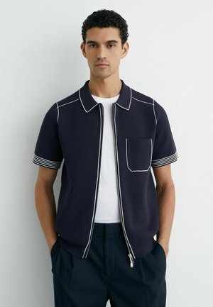 Mladý muž s krátkymi kučeravými vlasmi, oblečený v námornícky modrej krátky rukáv so zapínaním na zips cez bielu košeľu, stojaci pred jednoduchým bielym pozadím.