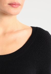 Musta neulepusero, jossa on leveä pääntie ja reunoissa resorikuvio. Kangas näyttää pehmeältä ja hieman joustavalta.