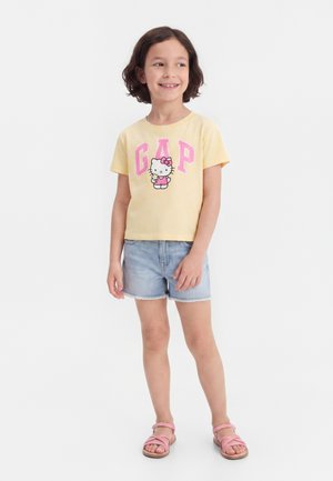 Χαμογελαστό παιδί με κοντά καστανά μαλλιά, που φοράει κίτρινο t-shirt Hello Kitty GAP, ανοιχτόχρωμα τζιν σορτς και ροζ σανδάλια, στέκεται μπροστά σε λευκό φόντο.