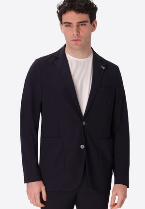 Uomo che indossa una giacca nera a due bottoni sopra una maglietta bianca con collo rotondo, in piedi davanti a uno sfondo chiaro uniforme.