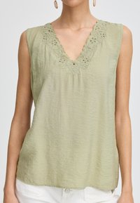 Top senza maniche di colore verde chiaro con scollatura a V, caratterizzato da ricami in pizzo decorativi intorno al collo e un tessuto testurizzato.