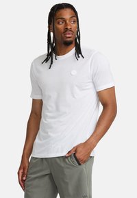 Vit kortärmad t-shirt med rund halsringning och en liten, cirkulär logotyp på bröstet. Slät tyg, avslappnad passform, minimalistisk design.