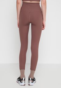 Magas derekú barna leggings sima textúrával, testhezálló kialakítással és hangsúlyos varrás részletekkel, fekete-fehér sportcipőkkel párosítva.