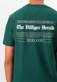 Grøn bomulds-T-shirt med et stort trykt tekstdesign på ryggen i hvid, med dekorative elementer og en vintage stil.