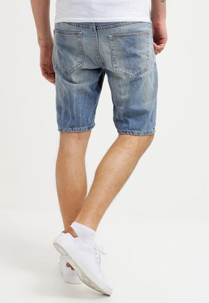 Hellblaue Jeansshorts mit einer lockeren Passform, umgeschlagenen Säumen und einem Fünf-Taschen-Design. Getragen mit weißen Turnschuhen und Socken.
