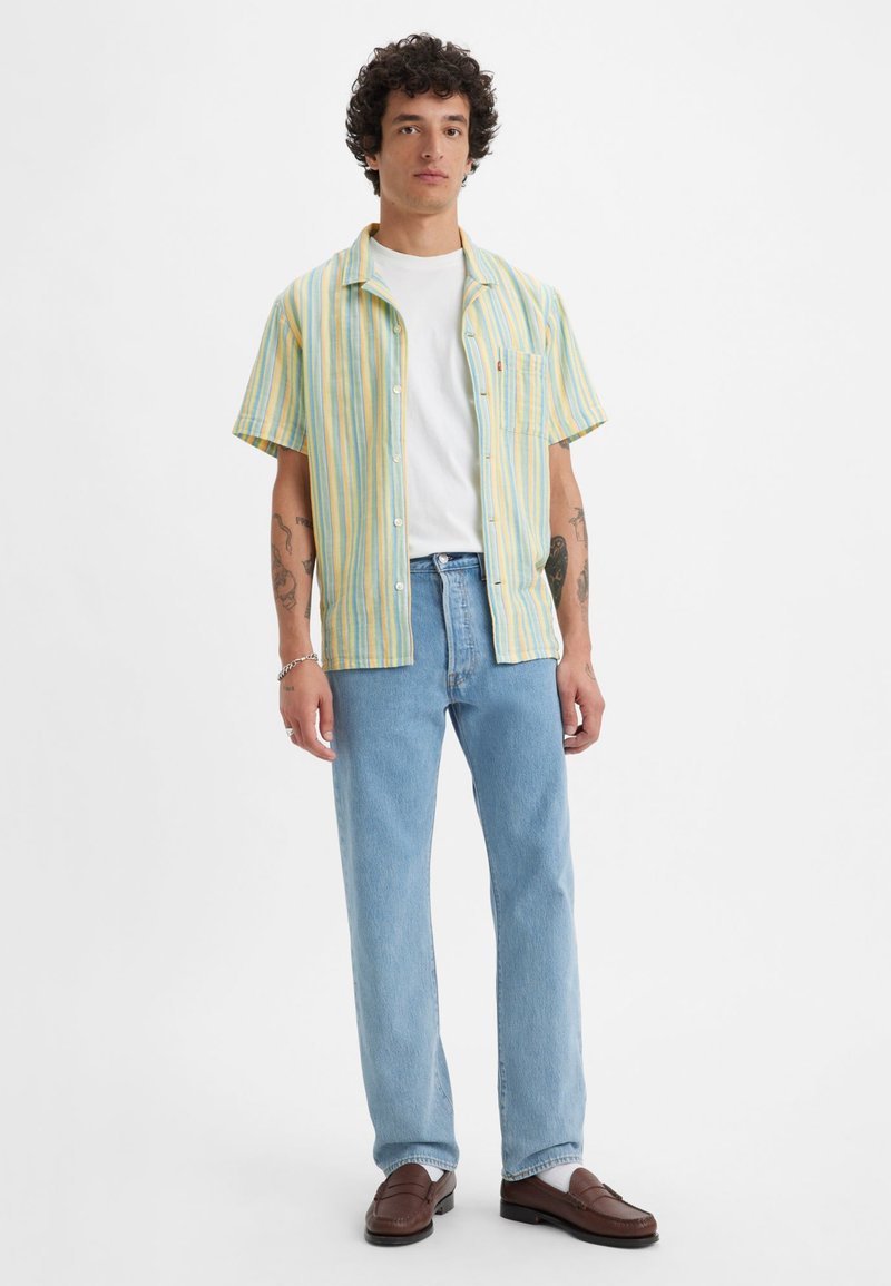Kurzarmhemd mit Knöpfen in Gelb- und Blaustreifen, kombiniert mit hellblauen Jeans und braunen Slippern. Darunter ein weißes T-Shirt.