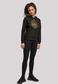 Zwarte hoodie met kleurrijke wapenafbeelding, voorzak, ribgebreide manchetten en zoom; gecombineerd met zwarte leggings en schoeisel.