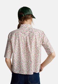 Camicetta a maniche corte in tessuto leggero, con uno sfondo bianco e un motivo floreale in rosa e verde. Dettaglio del colletto con bottoni.