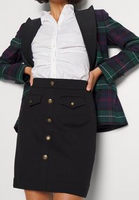 Svart blyertskjol med hög midja, frampocketar och detaljer i guldknappar. Bärs med en vit skjorta och en rutigt blazer.