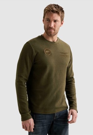 Homme portant une chemise à manches longues vert olive avec un col rond et des patchs brodés, associée à un jean bleu foncé, sur un fond uni.