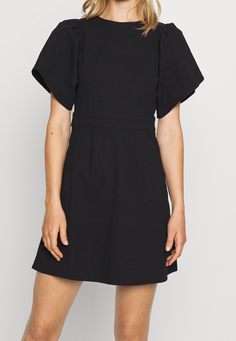 Femme portant une courte robe noire avec des manches courtes bouffantes et une taille cintrée, debout devant un fond clair uni.