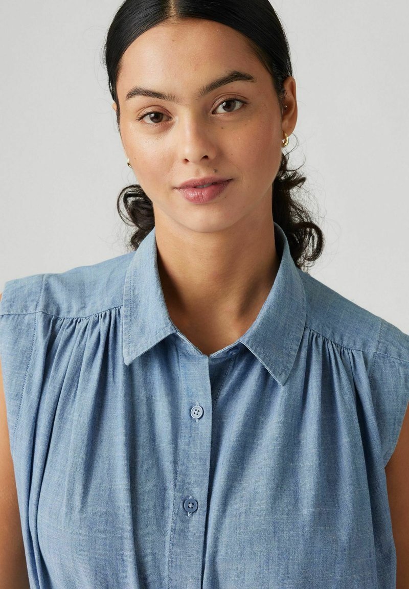 Chemise sans manches bleu clair avec un devant boutonné, un col et des détails froncés aux épaules, fabriquée à partir d'un tissu doux et texturé.