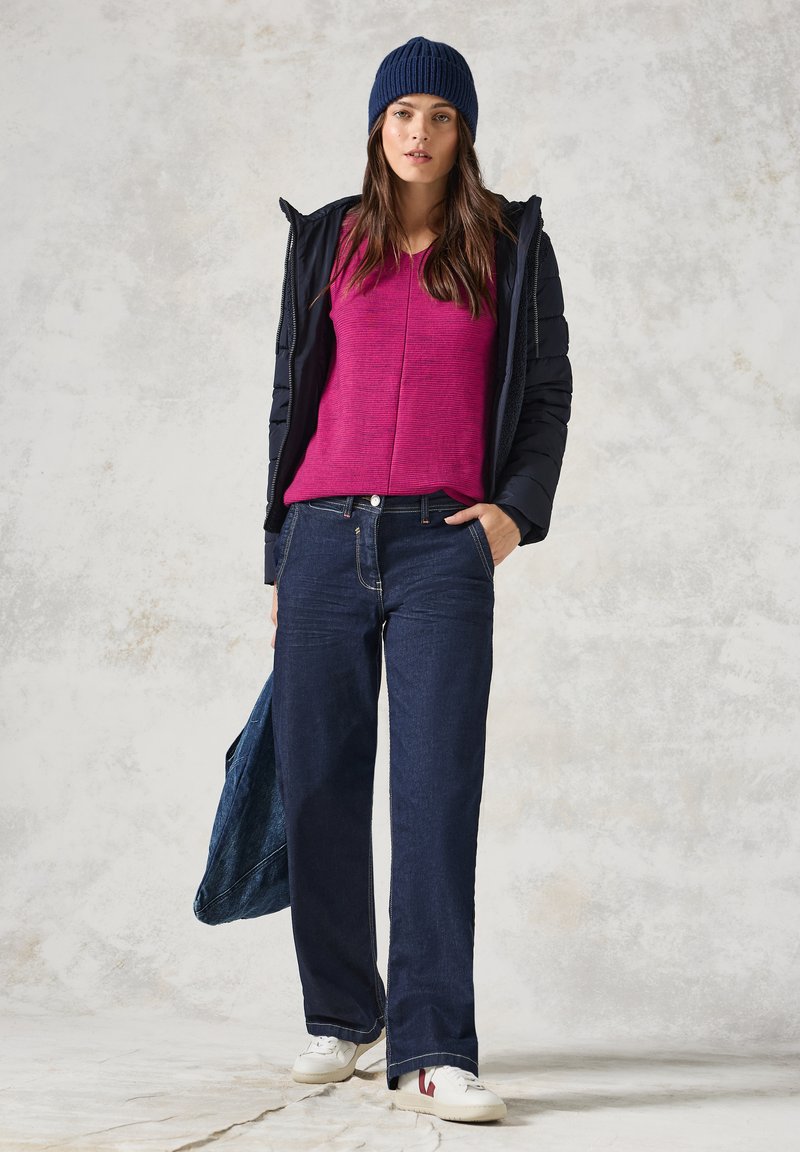 Frau, die eine marineblaue Daunenjacke, ein fuchsiafarbenes Oberteil und weit geschnittene, dunkle Jeans trägt, dazu eine marineblaue Strickmütze und weiße Sneakers.