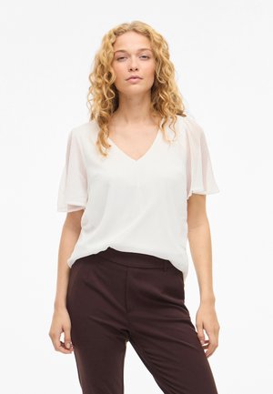 Sieviete ar cirtainiem blondiem matiem, tērpusies baltā V veida izgriezuma blūzē ar plandošām piedurknēm un tumši brūnās biksēs, stāv balta fona priekšā.