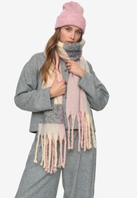 Donna che indossa un completo in maglia grigio, un berretto rosa e una lunga sciarpa a righe nei colori pastello rosa, crema e grigio con frange, su sfondo bianco.