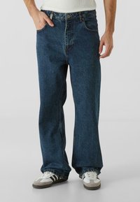 Blå denimjeans med en avslappet passform, som har sidelommer, knappfeste foran, og en subtil falmet tekstur. Kombinert med hvite og grå sneakers.