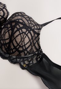 Soutien-gorge en dentelle noire avec motifs géométriques, bonnets transparents, bretelle réglable et petit détail logo doré sur le bandeau.