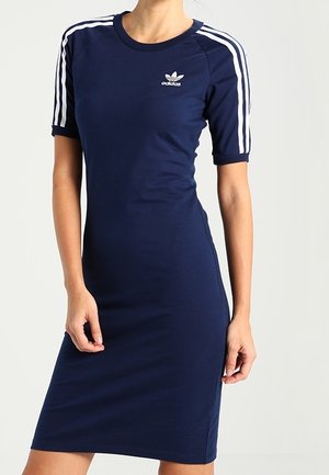 Femme portant une robe Adidas bleu marine ajustée avec des bandes blanches sur les manches courtes et le logo Adidas sur la poitrine.