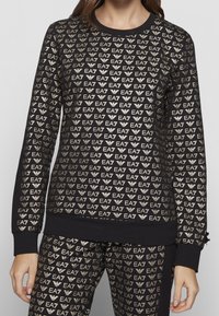 Svart sweatshirts med guld EA7-logomönster över hela, ribbad halsringning och ärmslut. Mjukt tyg med avslappnad passform och långa ärmar.