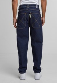 Pantalones vaqueros de mezclilla azul oscuro con un ajuste relajado, con dos bolsillos traseros con costuras amarillas y una etiqueta de marca en la cintura.