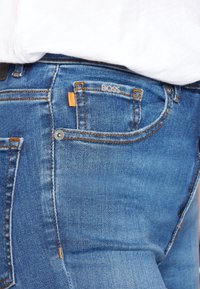 Blue denim jeans with orange accent at the pocket, silver hardware, and visible stitching. Features a logo on the back pocket.
