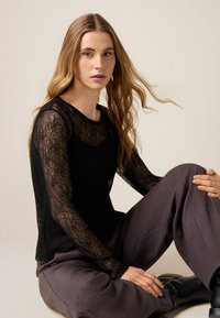 Top de encaje negro de manga larga con escote redondeado, combinado con pantalones grises de pierna ancha y botas negras hasta el tobillo. Textura suave y translúcida.