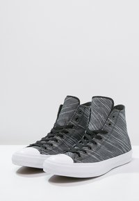 Zapatillas altas en blanco y negro con parte superior de textil a rayas, puntera de goma blanca y cordones negros. Suela plana con borde redondeado.